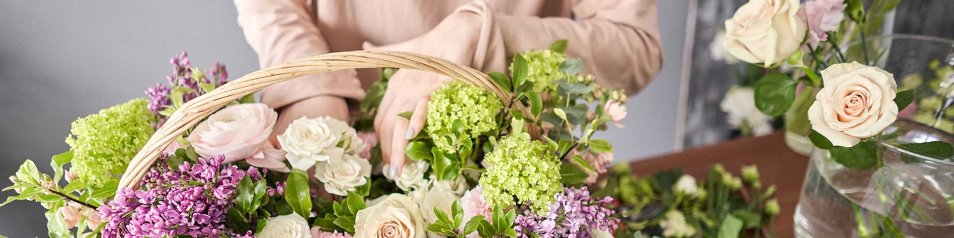 En person holder en kurv fyldt med farverige blomster, herunder roser, ranunkler og liljekonvaller. Der er også flere friske blomster på bordet, og stemningen er lys og indbydende. 